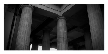 Black and white architectural photograph of massive classical columns supporting a grand structure, with dramatic high-contrast lighting.