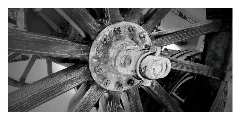 Black and white close-up photograph of a large wooden spoked wheel hub with visible bolts and aged wood texture.