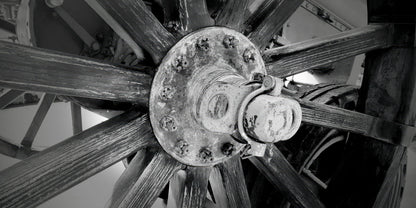 Black and white close-up photograph of a rustic wooden spoked wheel with a central metal hub and bolts.