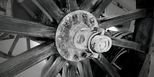 Black and white close-up photograph of a rustic wooden spoked wheel with a central metal hub and bolts.