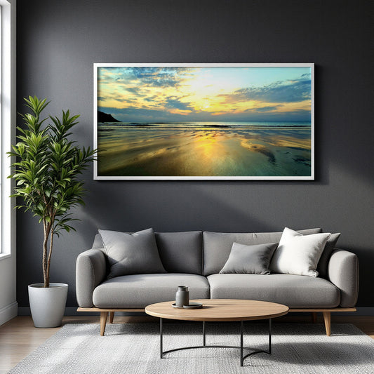 A large framed horizontal canvas print of a beach sunset with golden light reflecting on wet sand and blue-green water, displayed on a dark gray wall above a modern gray sofa in a styled living room.
