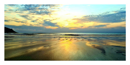 A scenic landscape photograph of a beach at sunset, showing golden light reflecting on wet sand and calm water, with a dark landform on the left and clouds in the sky.