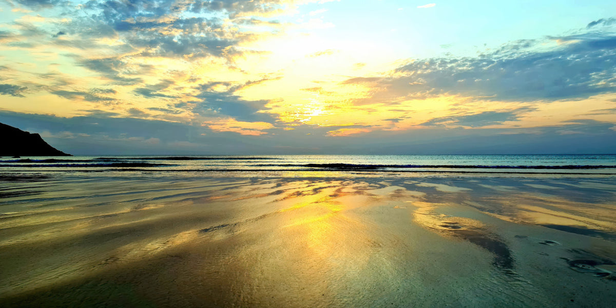 A scenic beach landscape at sunset with golden light reflecting on wet sand, calm ocean water, and a cloudy sky.