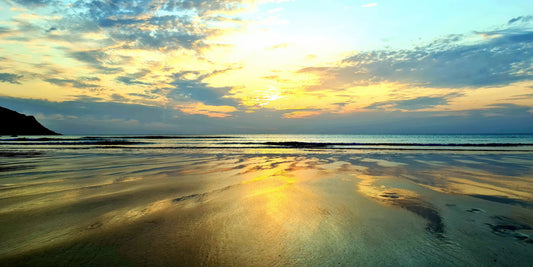 A scenic beach landscape at sunset with golden light reflecting on wet sand, calm ocean water, and a cloudy sky.
