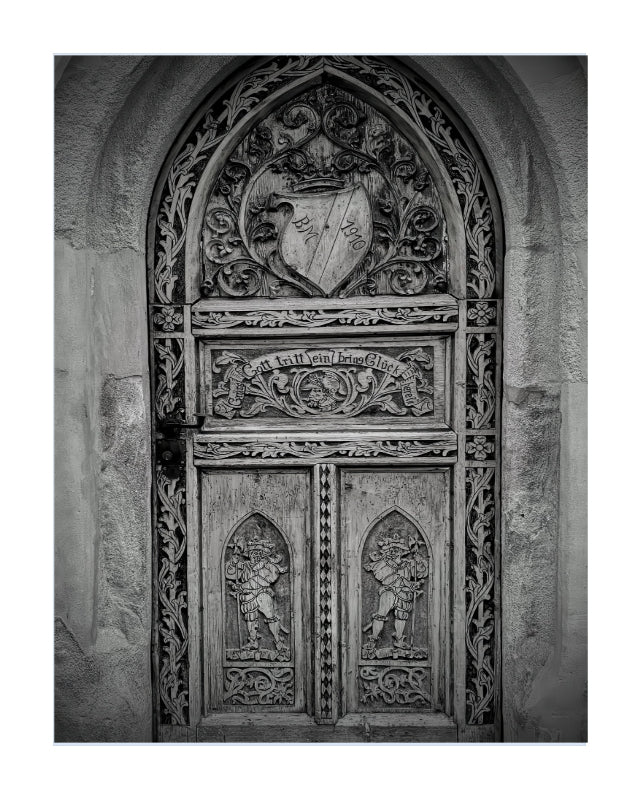 A black and white photograph of an ornate medieval-style carved wooden door set within a stone archway, presented as a square art print with a subtle drop shadow on a white background.