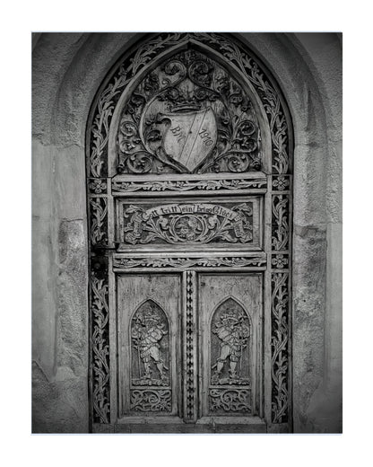 A black and white photograph of an ornate medieval-style carved wooden door set within a stone archway, presented as a square art print with a subtle drop shadow on a white background.
