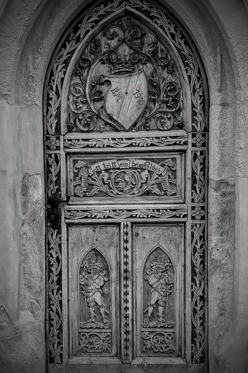 A detailed black and white photograph of an ornate, intricately carved wooden gothic door with a heraldic shield crest at the top.