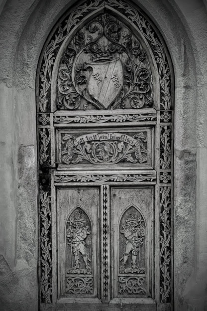 A detailed black and white photograph of an ornate, intricately carved wooden gothic door with a heraldic shield crest at the top.