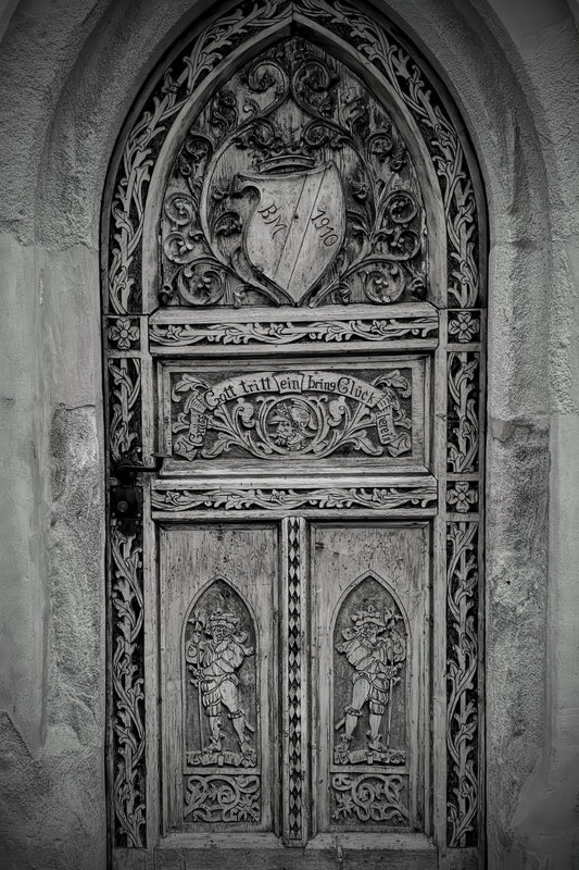 A detailed black and white photograph of an ornate, intricately carved wooden gothic door with a heraldic shield crest at the top.