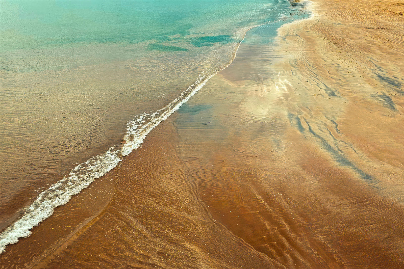 Aerial view of a sandy beach shoreline with turquoise water, white surf, and warm tan sand showing ripple patterns.