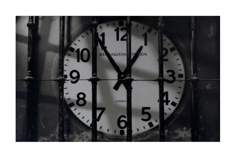 A square black-and-white photograph showing a London Underground Keinghamton Station analog clock viewed through vertical metal bars, with a white border around the artwork.