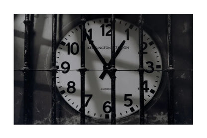 A square black-and-white photograph showing a London Underground Keinghamton Station analog clock viewed through vertical metal bars, with a white border around the artwork.