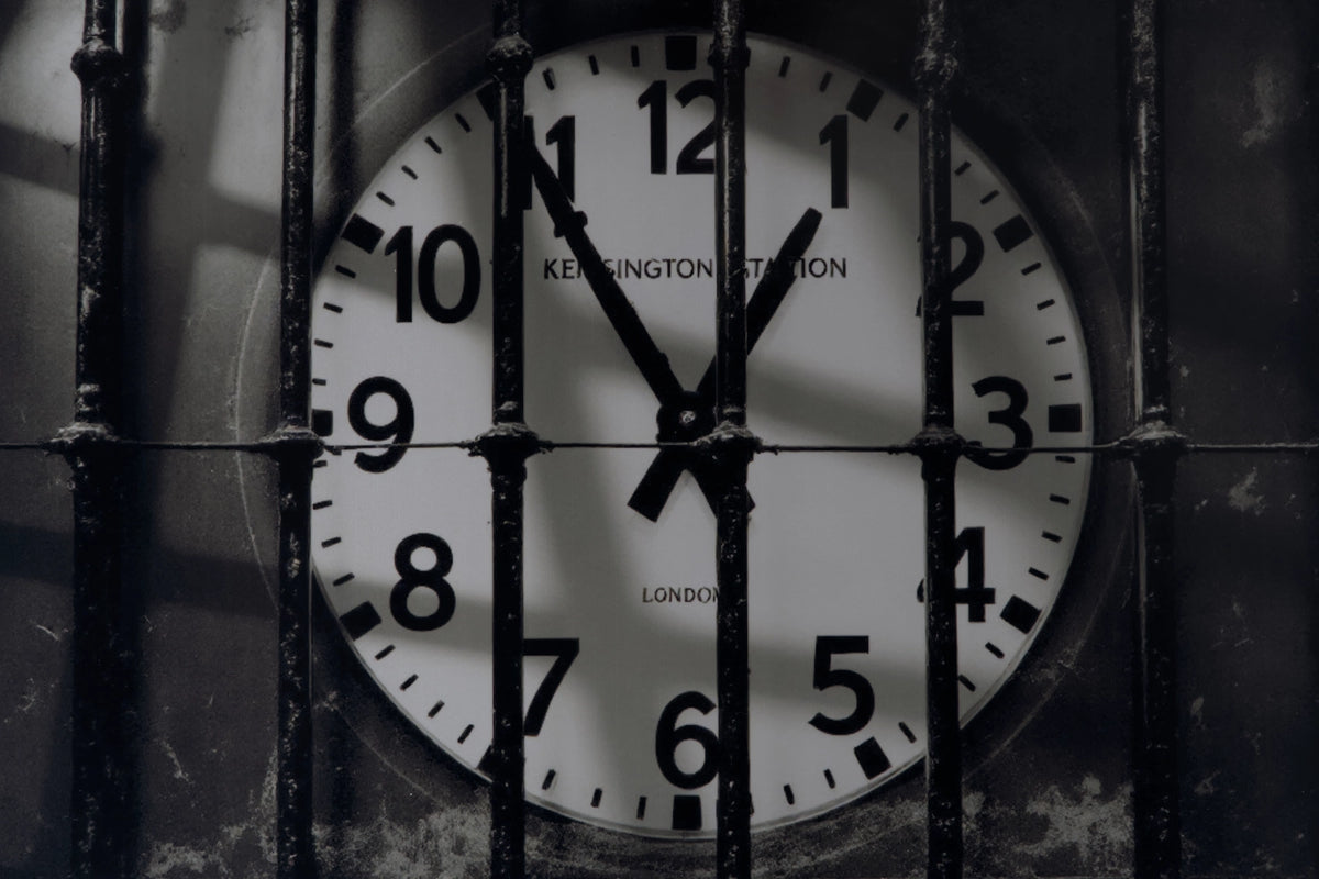 A close-up of a round wall clock showing 11:55, viewed through vertical metal bars. The clock face is white with black numerals and hands, and the words 'KENSINGTON STATION' and 'LONDON' are visible on the clock face.
