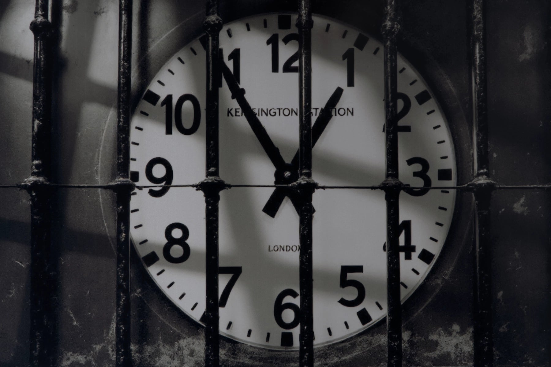 A close-up of a round wall clock showing 11:55, viewed through vertical metal bars. The clock face is white with black numerals and hands, and the words 'KENSINGTON STATION' and 'LONDON' are visible on the clock face.