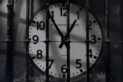 A close-up of a round wall clock showing 11:55, viewed through vertical metal bars. The clock face is white with black numerals and hands, and the words 'KENSINGTON STATION' and 'LONDON' are visible on the clock face.
