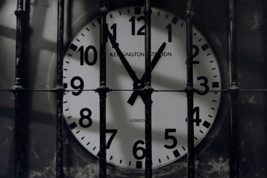 A close-up of a round wall clock showing 11:55, viewed through vertical metal bars. The clock face is white with black numerals and hands, and the words 'KENSINGTON STATION' and 'LONDON' are visible on the clock face.