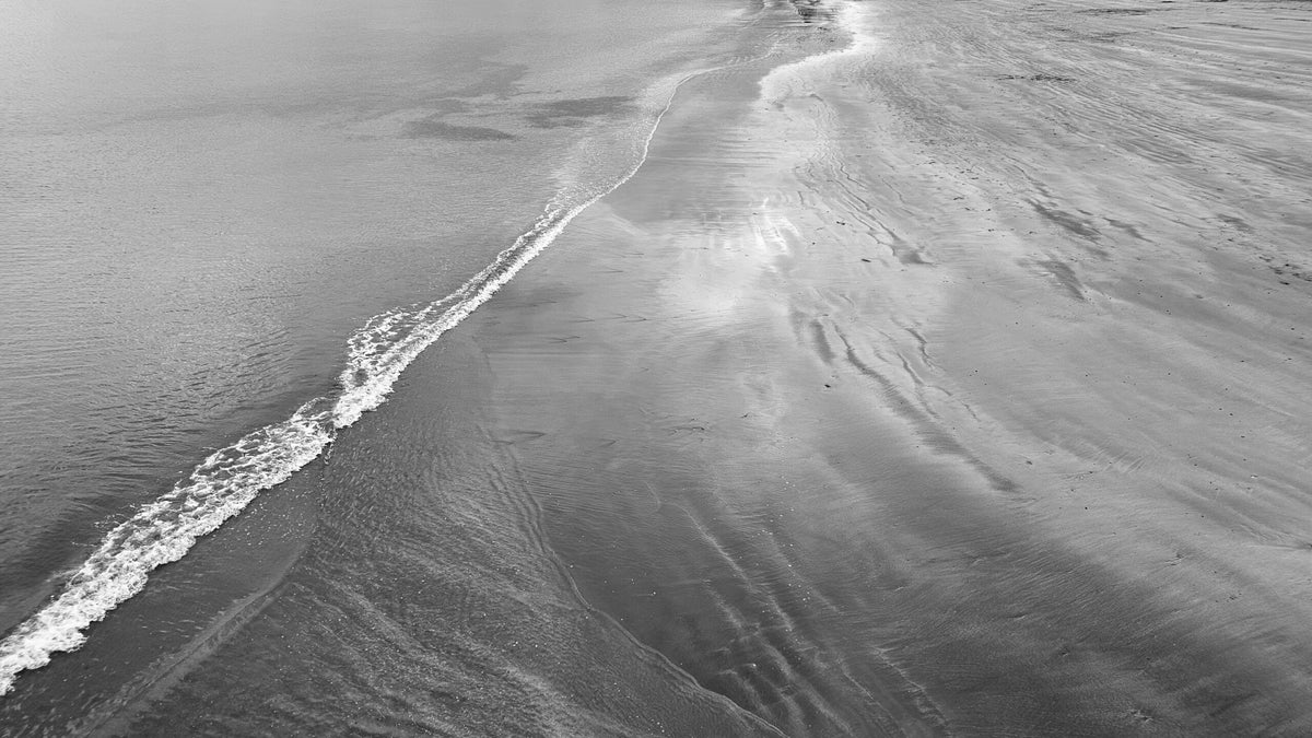 Black and white aerial photograph of a sandy beach with a shoreline curve, showing textured sand ripples and gentle waves.