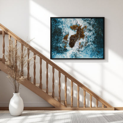 A framed wall art print showing an aerial view of blue ocean waves crashing around rocky shoreline, displayed on a white wall above a wooden staircase in a modern home interior.