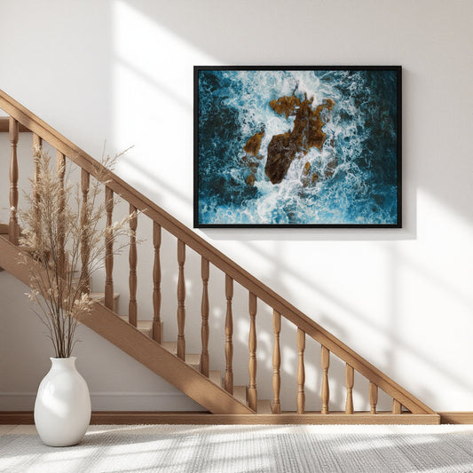 A framed wall art print showing an aerial view of blue ocean waves crashing around rocky shoreline, displayed on a white wall above a wooden staircase in a modern home interior.