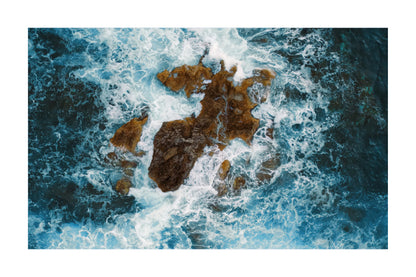 An aerial photograph of deep blue ocean waves crashing around dark brown rocky shoreline, presented as a square art print with a white border.