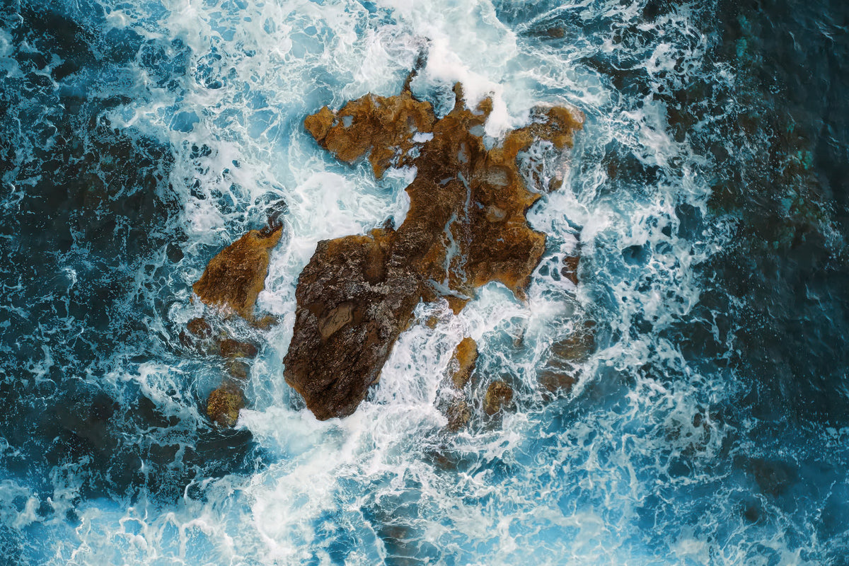 Aerial view of dark brown rocks surrounded by churning blue ocean water with white sea foam.