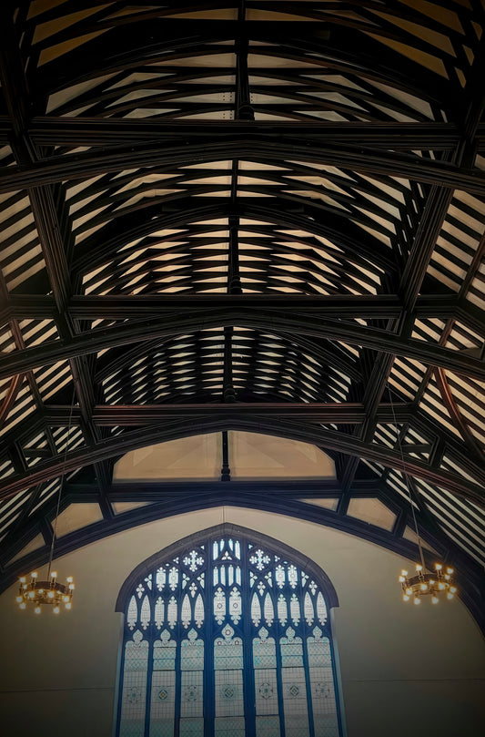 An upward view of a large arched cathedral-style ceiling with dark wooden beams and a tall stained-glass window at the bottom, with chandeliers visible on both sides.
