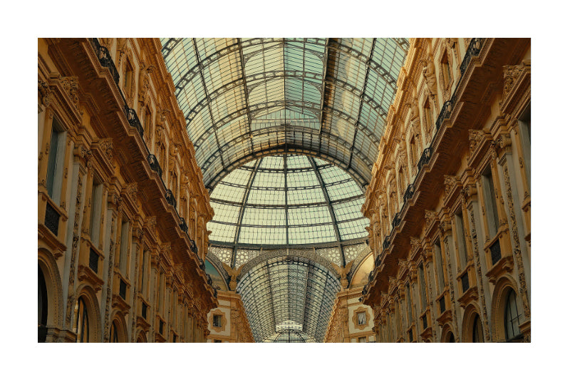 An architectural photograph of a grand glass-domed gallery with ornate beige and gold-toned stone columns on both sides, shown centered on a white background.