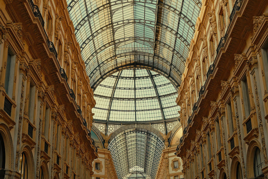 The grand arched glass and metal roof of the Galleria Vittorio Emanuele in Milan, overlooking ornate beige stone arcade buildings on both sides.