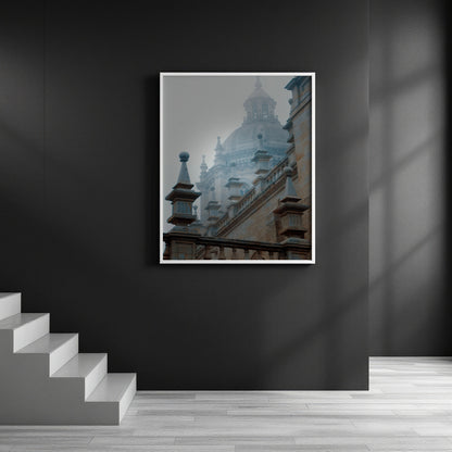 A framed art print depicting a misty, foggy architectural scene of a historic cathedral and building facades, displayed on a dark gray wall in a modern interior setting with a staircase on the left.
