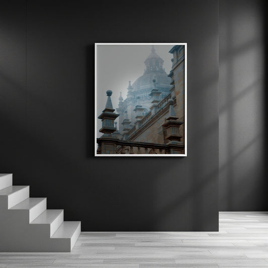 A framed art print depicting a misty, foggy architectural scene of a historic cathedral and building facades, displayed on a dark gray wall in a modern interior setting with a staircase on the left.
