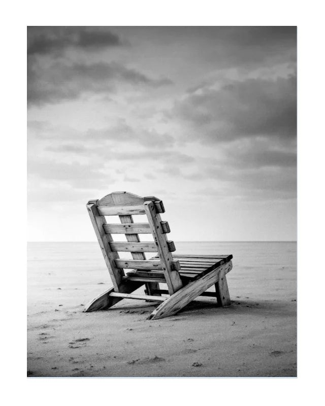 Black and white photograph of a wooden beach chair placed on a sandy shore under a cloudy sky.