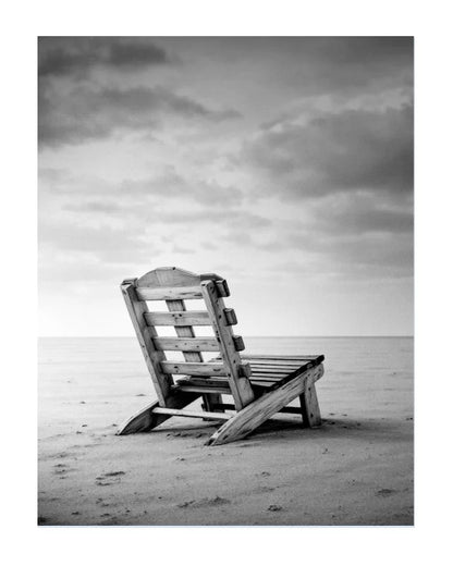 Black and white photograph of a wooden beach chair placed on a sandy shore under a cloudy sky.