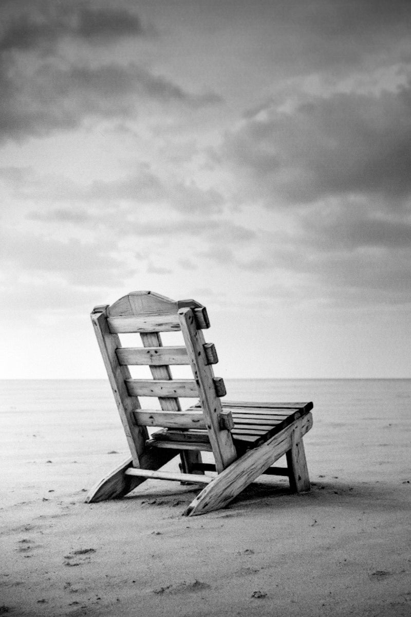 A weathered wooden beach chair sits on a sandy shore facing a calm ocean under a cloudy sky, shown in a black and white photograph.