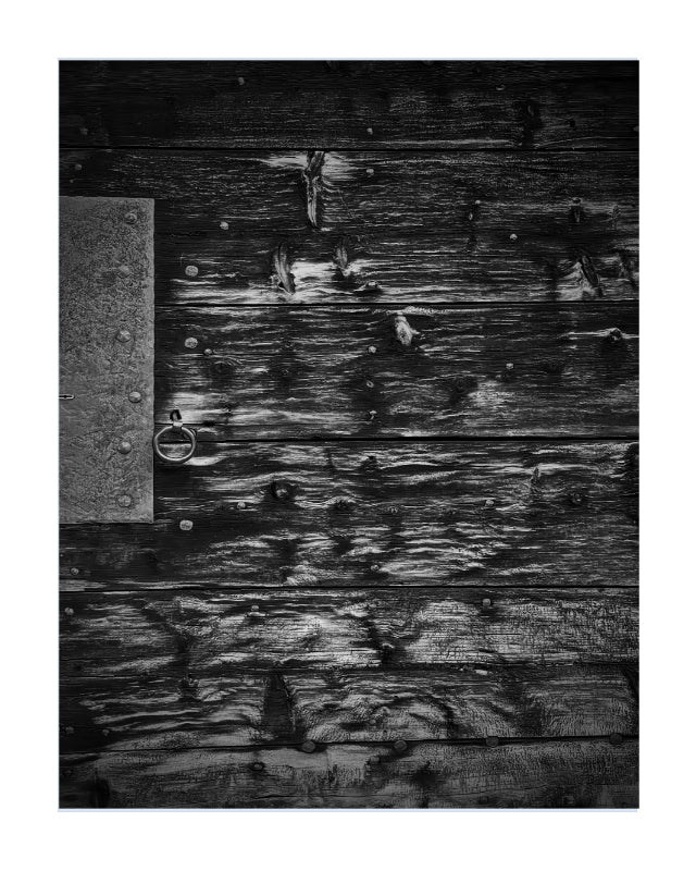 A black and white photograph of weathered wooden planks with a metal door knocker on the left side, presented as a square art print with a white border.
