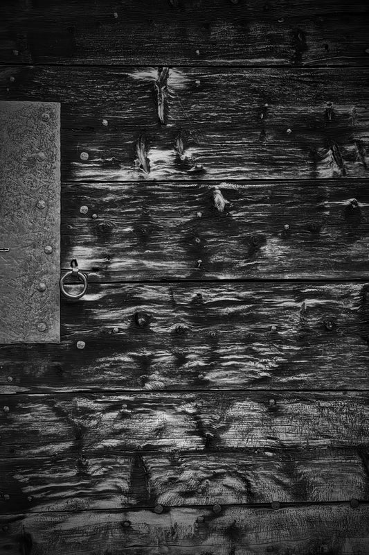 Black and white close-up of weathered wooden planks with a metal hinge and ring pull on the left side.
