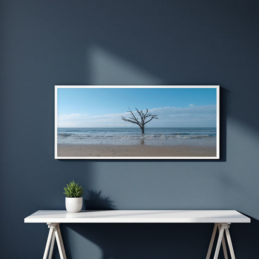A framed panoramic landscape print of a lone tree on a beach under a blue sky, displayed on a dark blue wall above a white console table with a small potted plant.