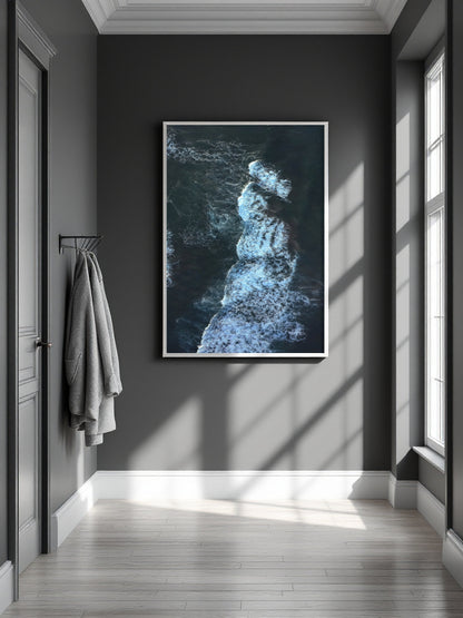 A framed vertical art print depicting a dark blue and white ocean wave crashing, displayed on a dark gray wall in a modern hallway with light wood floors and a hanging gray towel.
