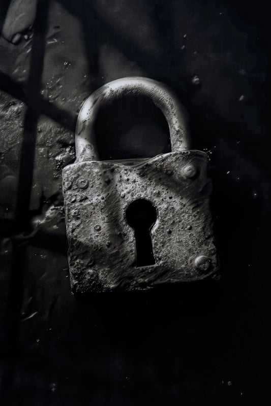 A weathered, textured metal padlock with a keyhole cutout, photographed in dramatic low light against a dark background with shadowy window-like lines.