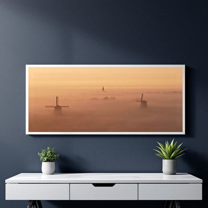 A framed landscape art print depicting windmills rising above a misty orange and yellow foggy horizon, displayed on a dark blue wall above a white console table with two small potted plants.