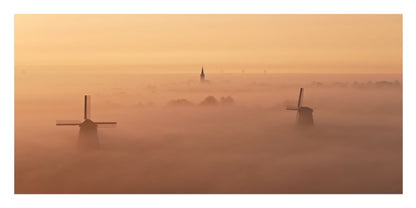 A misty landscape print featuring two dark windmills rising above a thick fog bank under a warm golden-orange sky.