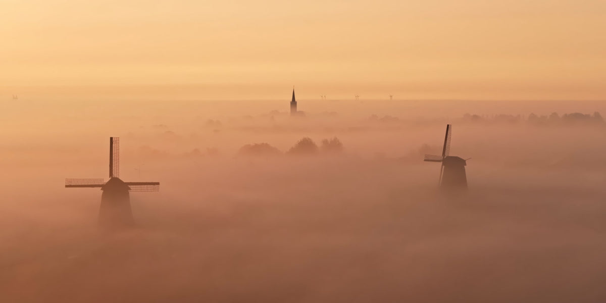 A misty landscape at sunrise or sunset with two windmills standing above thick fog, and a church spire faintly visible in the distance under a warm orange sky.