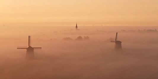 A misty landscape at sunrise or sunset with two windmills standing above thick fog, and a church spire faintly visible in the distance under a warm orange sky.