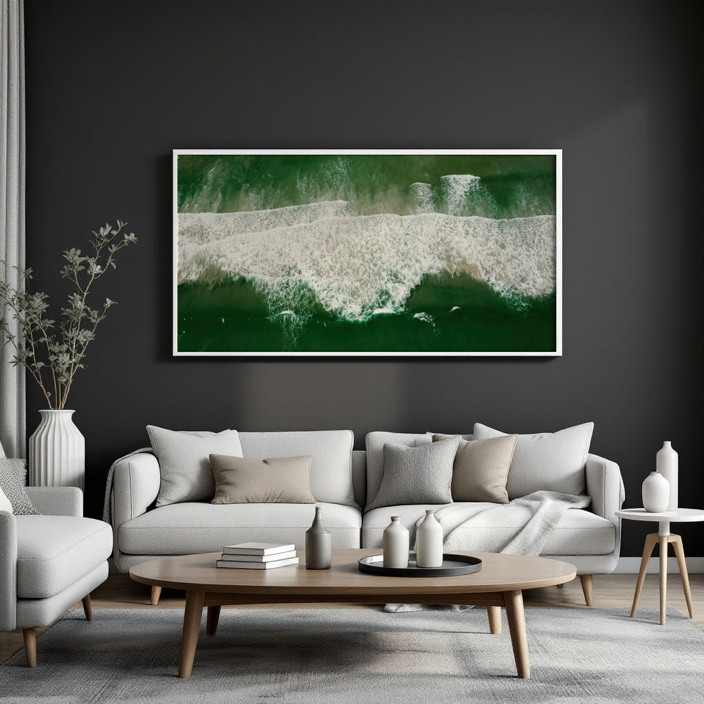 A framed horizontal canvas print of an aerial ocean wave scene in deep green and white, displayed on a dark charcoal wall above a modern light gray sofa in a styled living room.
