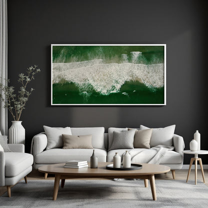 A framed horizontal canvas print of an aerial ocean wave scene in deep green and white, displayed on a dark charcoal wall above a modern light gray sofa in a styled living room.