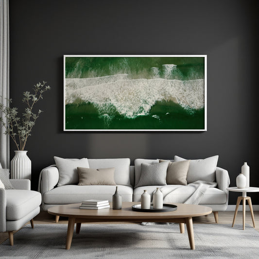 A framed horizontal canvas print of an aerial ocean wave scene in deep green and white, displayed on a dark charcoal wall above a modern light gray sofa in a styled living room.