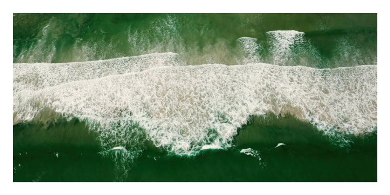 An aerial view of ocean waves rolling onto a shore, with deep green water and white sea foam.