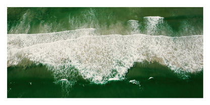 An aerial view of ocean waves rolling onto a shore, with deep green water and white sea foam.