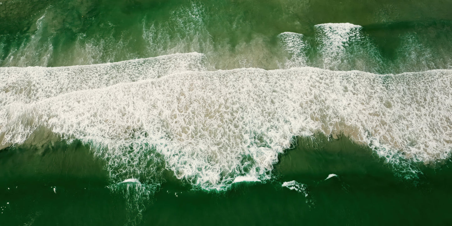Aerial view of green ocean waves with white foam rolling onto a sandy shore.