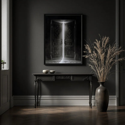 A framed black-and-white art print depicting an open door with light streaming in, displayed on a dark gray wall above a console table in a moody, dimly lit room with a large vase of dried grass nearby.