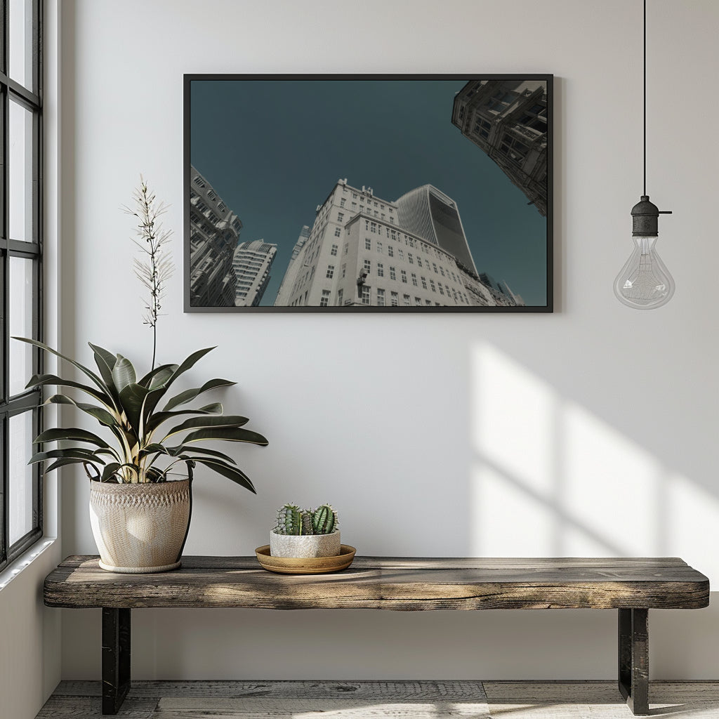 A framed wall art print depicting an upward-looking view of city skyscrapers under a dark teal sky, displayed on a white wall above a rustic wooden bench with a potted plant and a hanging light bulb in a modern room setting.